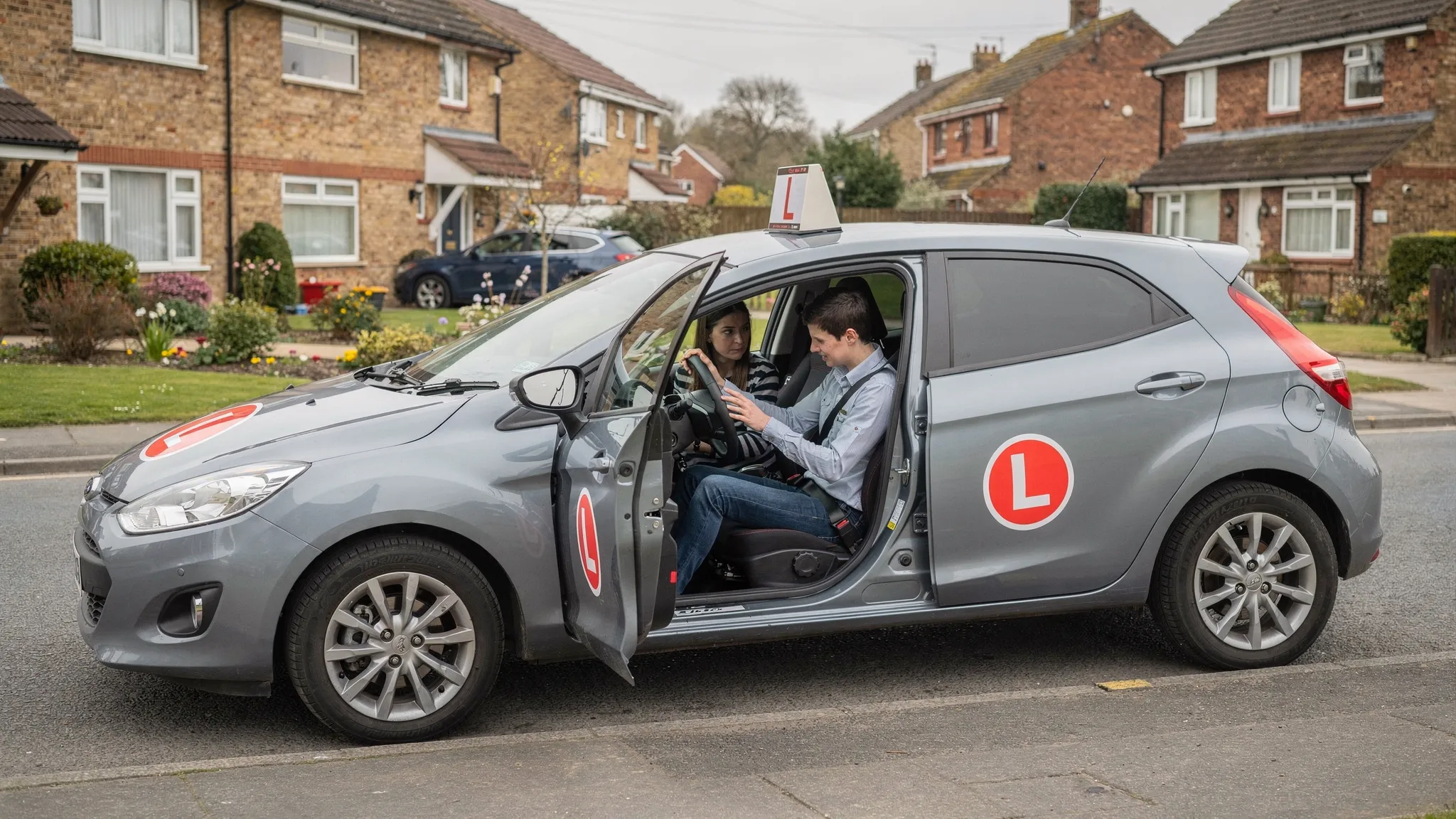 A learner car parked safely on a quiet residential road with L plates visible, while an instructor explains the basic controls to a first-time learner from the passenger seat.