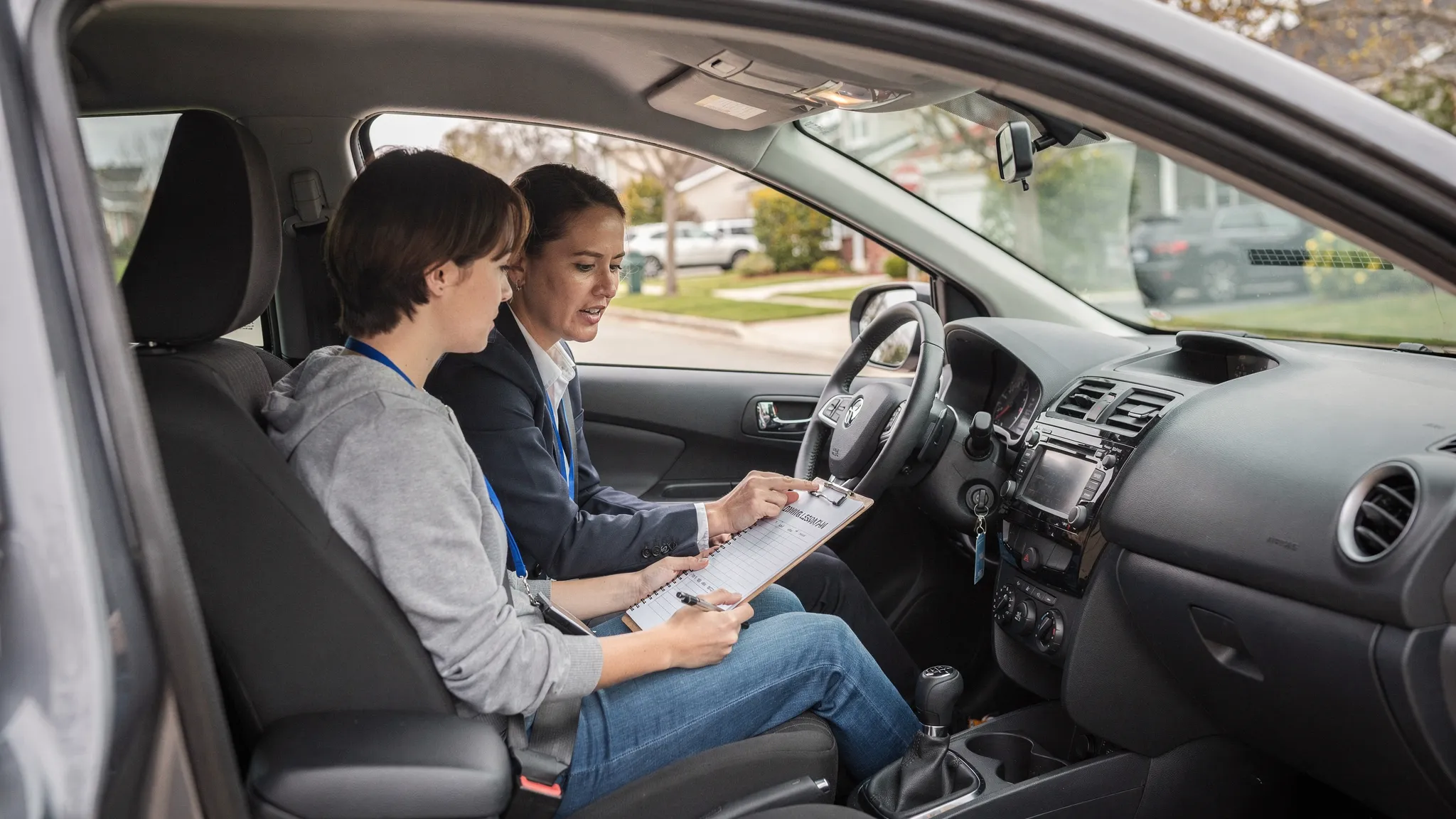 A learner driver and a driving instructor sitting in a parked dual-control car, reviewing a lesson plan together before setting off on a quiet residential road.