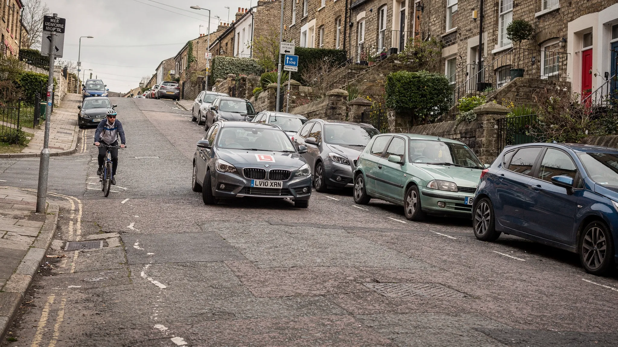 Street-level view of a learner car driving carefully up a steep Bristol residential street, with parked cars along the kerb, a cyclist passing and terraced houses rising on the slope.