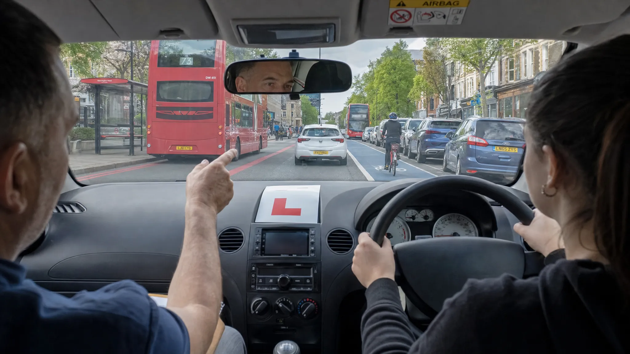 A learner car driving along a busy London street with buses, cycle lanes, parked cars and a driving instructor calmly guiding the learner from the passenger seat.