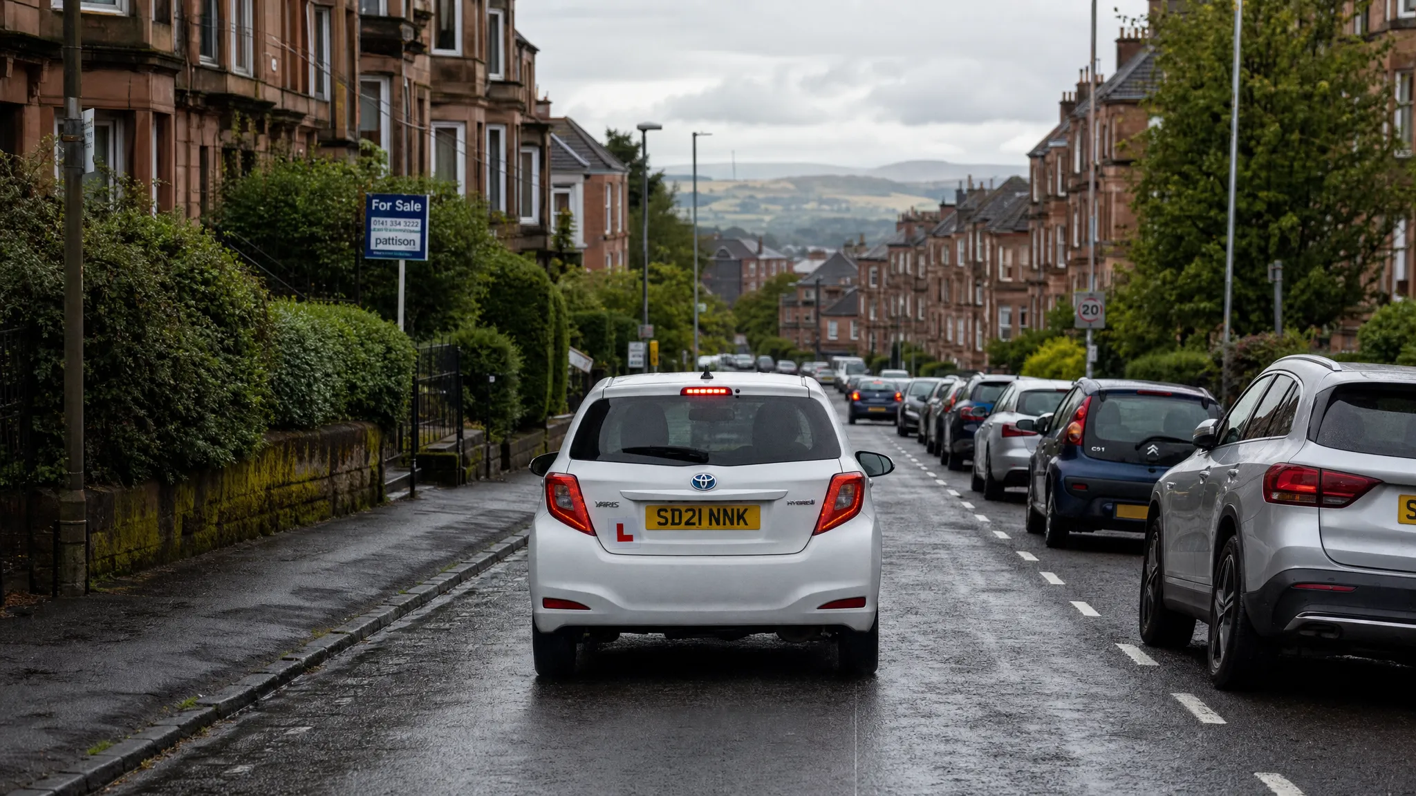 A learner car driving along a calm residential street in Glasgow with terraced houses, parked cars, light traffic and wet road conditions after rain.
