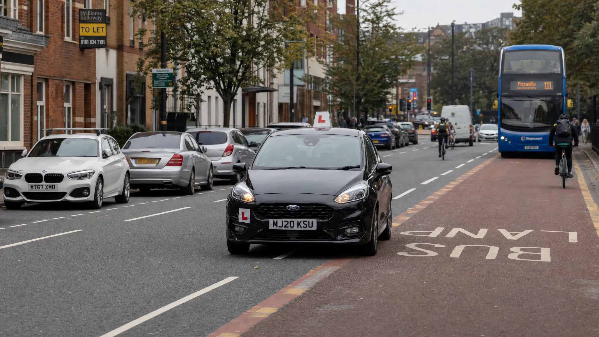 A learner car driving along a Manchester suburban street with parked cars, cyclists, bus lane markings and mixed city traffic in the background.