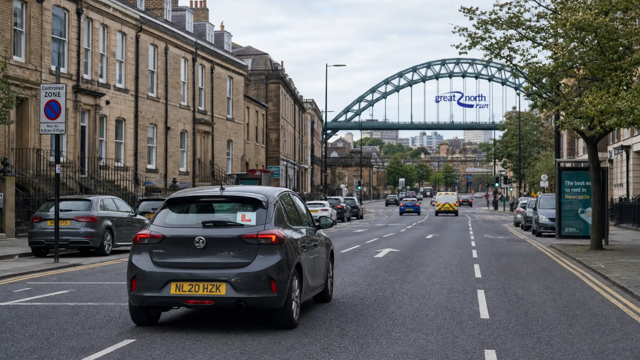 A learner car driving along a Newcastle street with terraced houses, light traffic, a visible L plate and the Tyne Bridge in the distance. The road scene shows clear lane markings, parked cars and a calm urban driving environment.