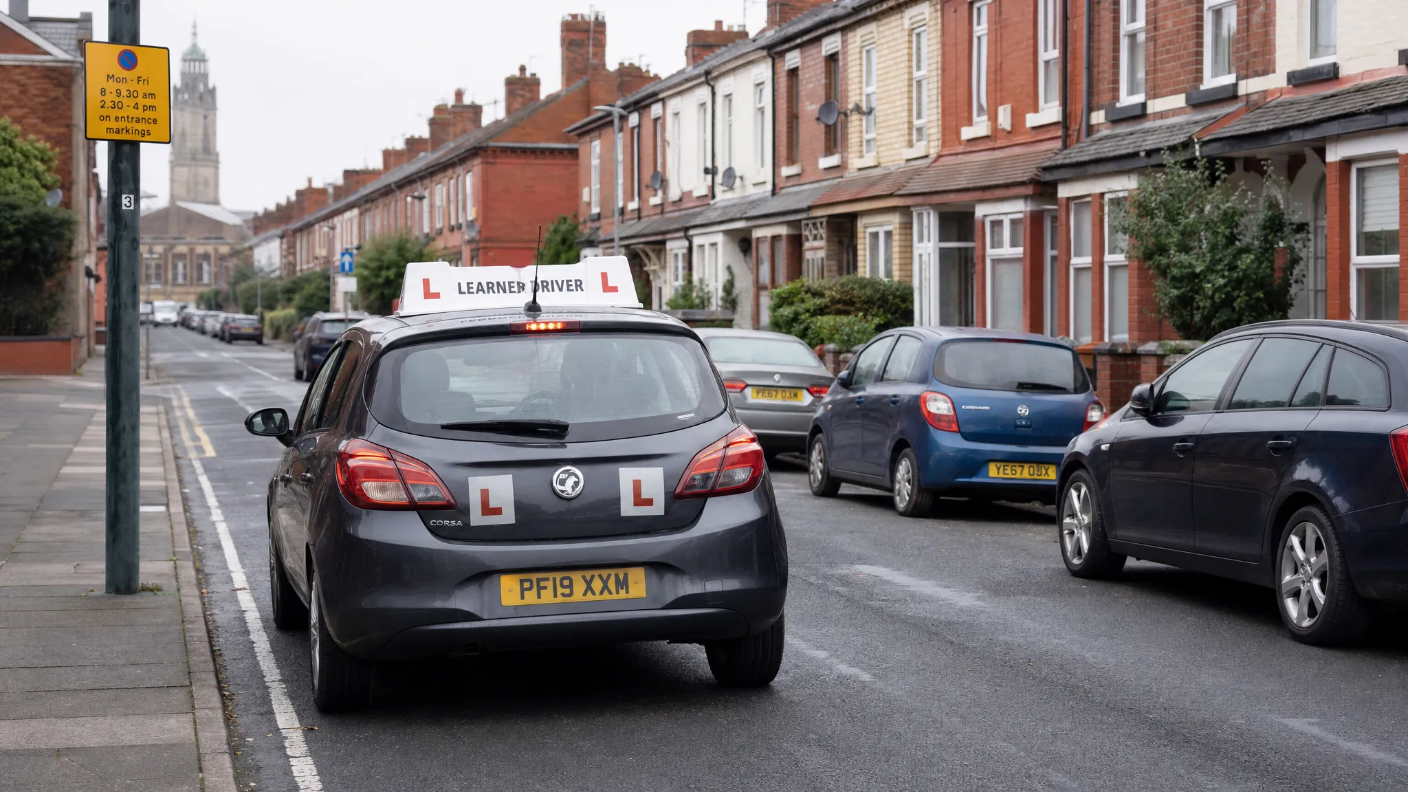 A learner car driving along a residential Liverpool street with terraced houses, parked cars and clear L plates, showing a calm driving lesson environment.