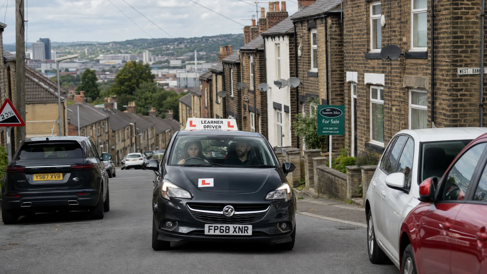 A learner car driving uphill on a Sheffield residential street with terraced houses, parked cars and clear L plates visible, while the instructor sits calmly in the passenger seat.