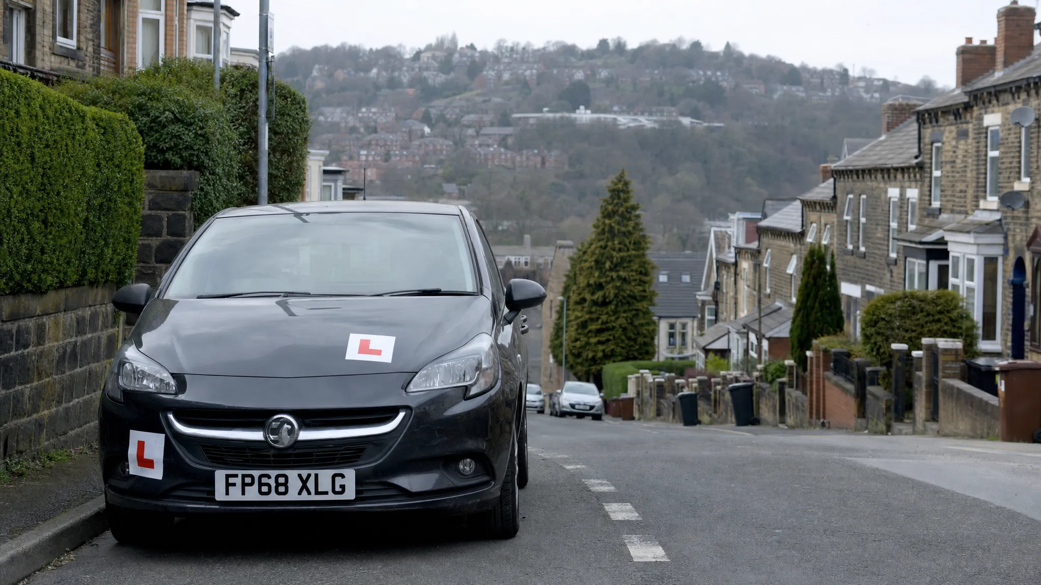 A learner car parked safely on a quiet Sheffield residential street with terraced houses and steep hills in the background, with L plates clearly visible.