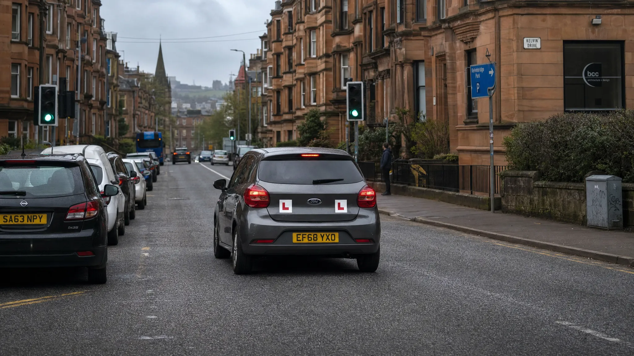 A learner car with L plates driving through a typical Glasgow street with parked cars, traffic lights and sandstone buildings in the background.